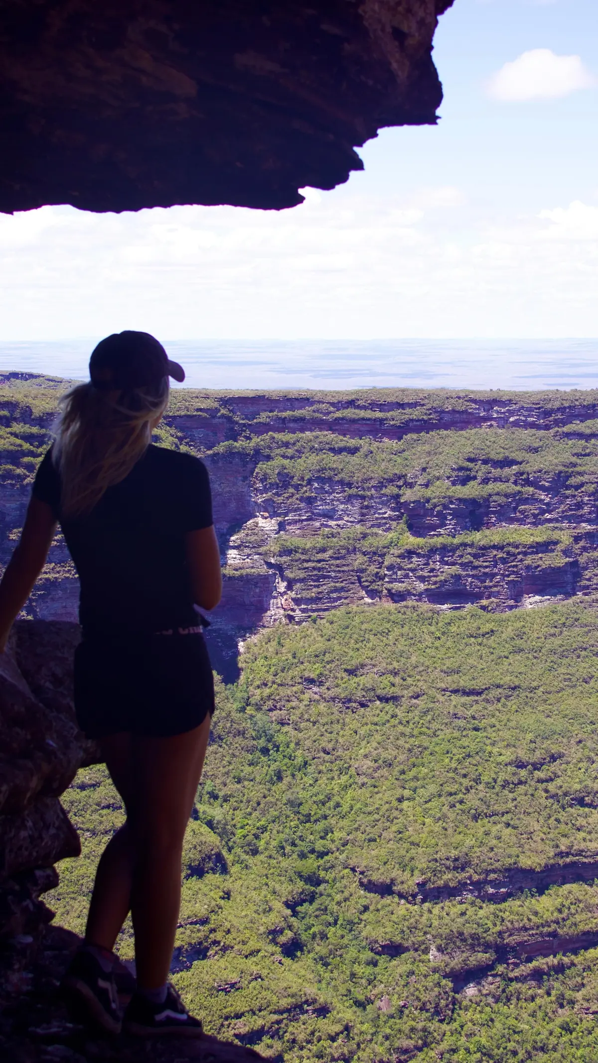 Mulher sob uma laje de pedra olhando um cânion enorme coberto de mata, com a planície no horizonte
