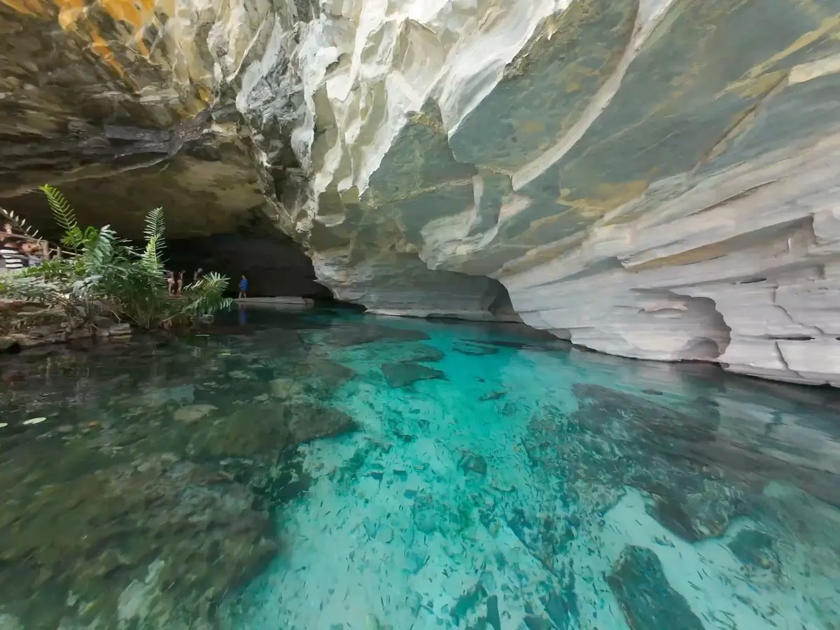 Gruta da Pratinha: água azul-turquesa transparente sob um paredão de rocha clara, com visitantes na entrada da gruta
